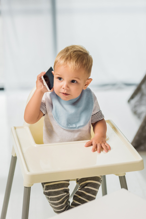 Cute toddler talking on smartphone and sitting in baby chairの写真素材