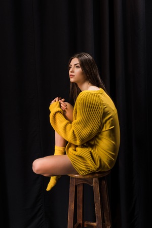 Side view of thoughtful woman in yellow woolen sweater sitting on bar stool on black backdropの写真素材
