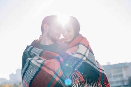 beautiful young couple covering in plaid and cuddling with sun shining behindの写真素材