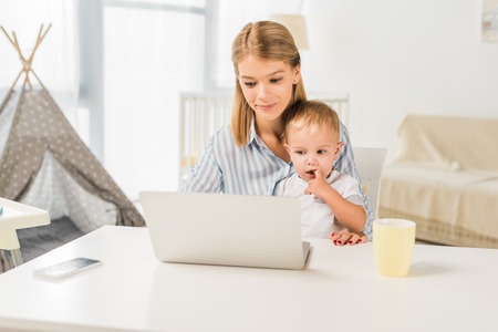 mother sitting at desk with cute toddler while using laptopの写真素材
