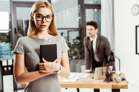 Selective focus of young female lawyer with notebook looking at camera while colleague standing at workplace in officeの写真素材
