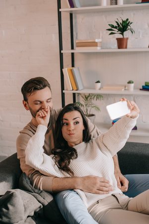 Playful young couple taking selfie with smartphone on couch at homeの写真素材