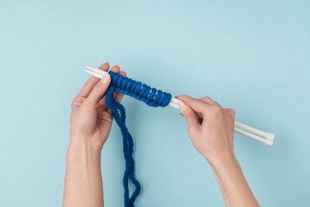 Partial view of woman with blue yarn and white knitting needles knitting on blue backdropの写真素材