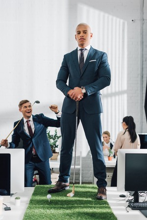 confident young african american businessman looking at camera while playing golf with colleague in officeの写真素材