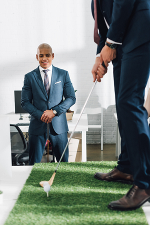 cropped shot of businessman playing golf and smiling african american colleague standing behind in officeの写真素材