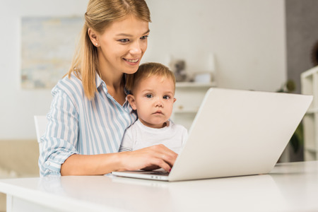Mother sitting at desk with cute toddler while using laptopの写真素材
