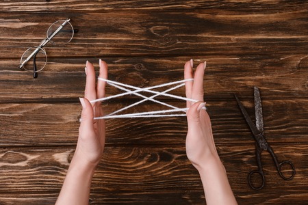 Partial view of woman holding white yarn for knitting on hands on wooden tabletop with eyeglasses and scissorsの写真素材
