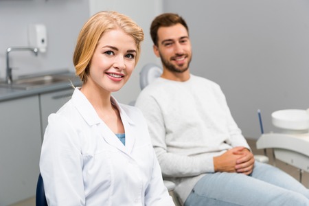 smiling female dentist and handsome young client looking at camera in dentist officeの写真素材