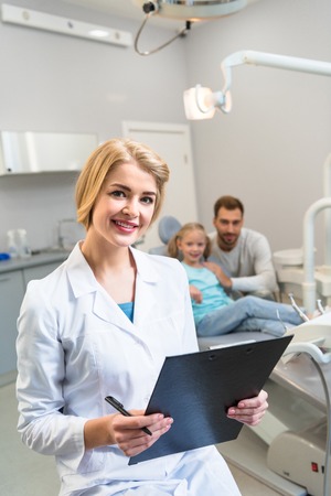 young female dentist with clipboard looking at camera while little child and her father sitting on backgroundの写真素材