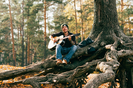adult male playing acoustic guitar sitting on tree in parkの写真素材