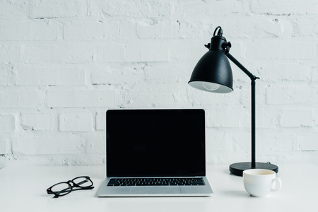 Laptop with blank screen, eyeglasses, lamp and cup on tableの写真素材