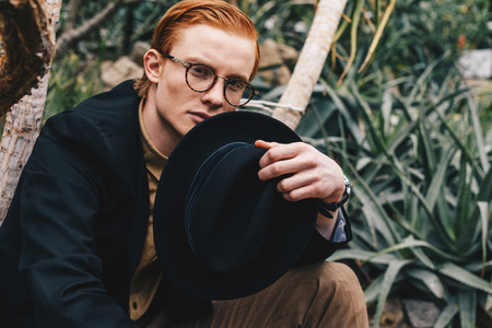 handsome young redhead man in eyeglasses holding hat and looking at cameraの写真素材