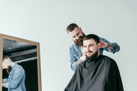 smiling barber shaving customer hair at barbershop isolated on whiteの写真素材