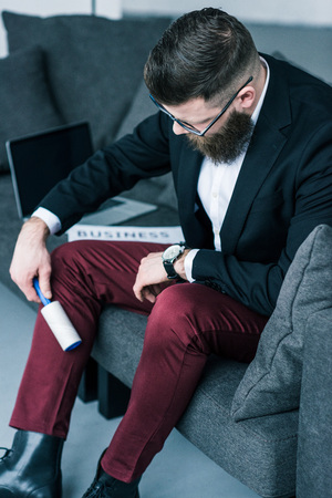 businessman cleaning pants with sticky brush while sitting on sofaの写真素材