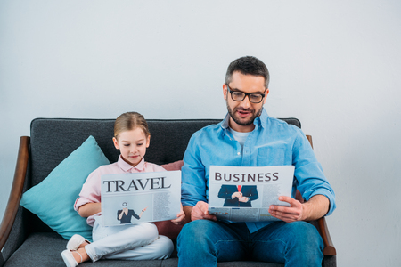 father and daughter reading newspapers while resting on sofa at homeの写真素材