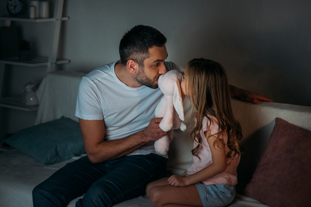 father and little daughter kissing toy while sitting on sofa at homeの写真素材