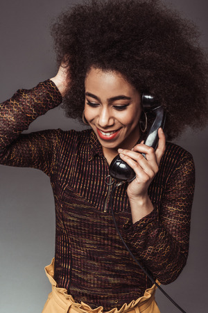 smiling african american retro styled girl talking by vintage stationary telephone isolated on greyの写真素材