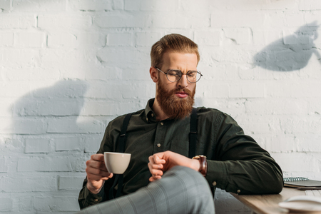 handsome bearded businessman holding cup of coffee and checking timeの写真素材