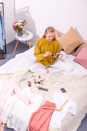 high angle view of young woman opening eye shadows box while sitting on bedの写真素材