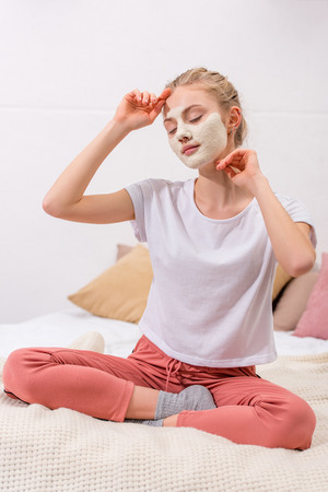 young woman with white clay facial mask sitting on bed at homeの写真素材