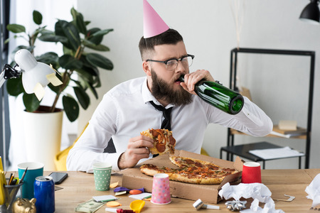 businessman with party cone on head drinking champagne while eating pizza at workplaceの写真素材