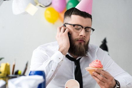 portrait of businessman in party cone with birthday cupcake in hand talking on smartphoneの写真素材