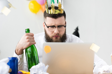 businessman with paper crown on head and bottle of champagne in hand looking at laptop screen at workplaceの写真素材