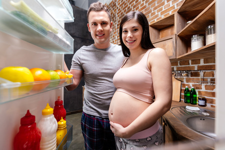 smiling young pregnant couple looking into refrigerator at kitchenの写真素材