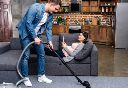 young man cleaning carpet with vacuum cleaner while pregnant woman lying with digital tablet on sofaの写真素材