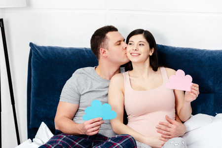 happy young pregnant couple holding blue and pink clouds symbols and kissing on bedの写真素材