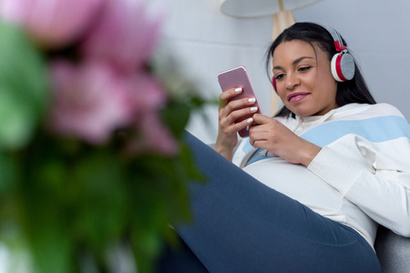 african american pregnant woman listening music with headphones and using smartphone