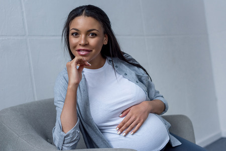 smiling african american pregnant woman looking at camera while sitting on armchairの写真素材