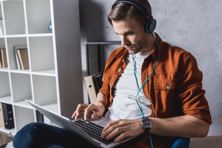 young handsome man in headphones working with laptop on couchの写真素材