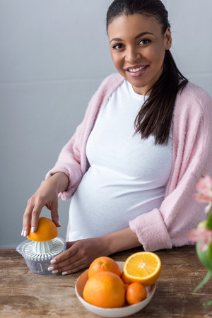beautiful smiling african american pregnant woman making orange juice with squeezerの写真素材