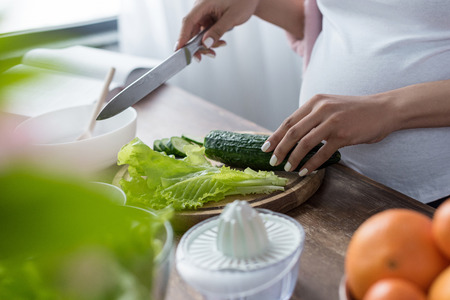 cropped view of pregnant woman cutting cucumber and salad at kitchenの写真素材