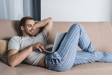 young relaxed freelancer in pajamas working with laptop on couchの写真素材