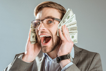 rich happy businessman holding dollar bills near his face isolated on greyの写真素材