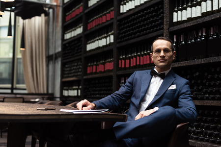 happy handsome man in stylish suit sitting in armchair in front of wine storage