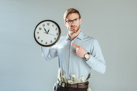 Young confident businessman pointing on clock isolated on grey