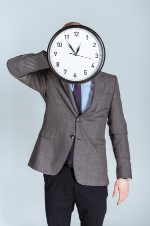 Businessman in suit holding clock over his face isolated on greyの写真素材