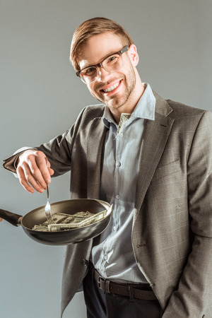 Young businessman frying dollar bills in pan isolated on greyの写真素材