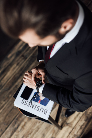overhead view of businessman looking at wristwatch while standing at stool with business newspaper and smartphoneの写真素材