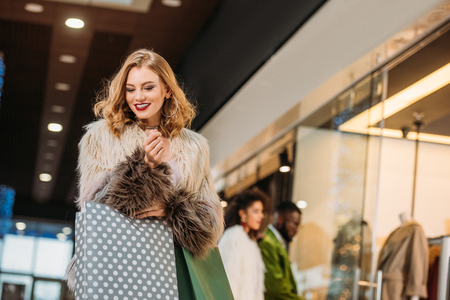 smiling fashionable young woman in fur coat holding shopping bags while walking in mallの写真素材