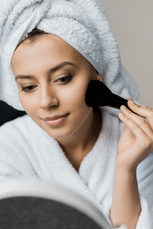 smiling woman in bathrobe and towel applying powder with cosmetic brushの写真素材
