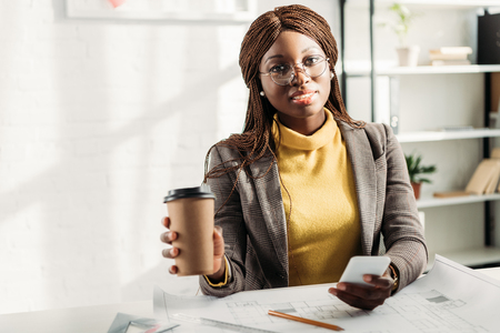 african american female architect in glasses holding coffee to go, using smartphone and working at desk on project with blueprintsの写真素材