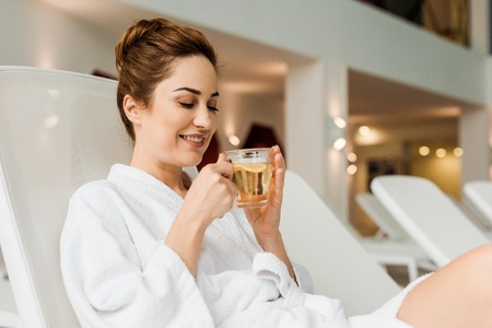 smiling young woman holding cup with herbal drink while relaxing in spa