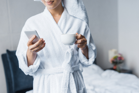 cropped view of woman in bathrobe holding cup of coffee and using smartphone in bedroom in the morningの写真素材