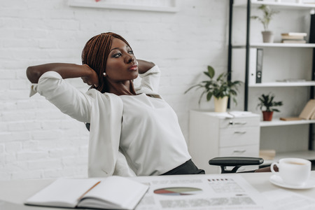 african american adult businesswoman in white formal wear with hands behind head sitting at desk and relaxing at modern officeの写真素材
