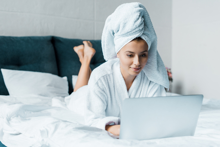 beautiful young woman in bathrobe using laptop while lying on bed in the morningの写真素材