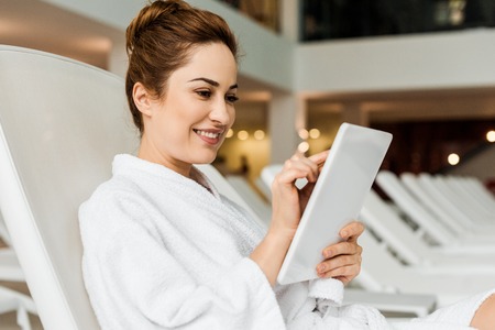smiling young woman in bathrobe using digital tablet while relaxing in spaの写真素材
